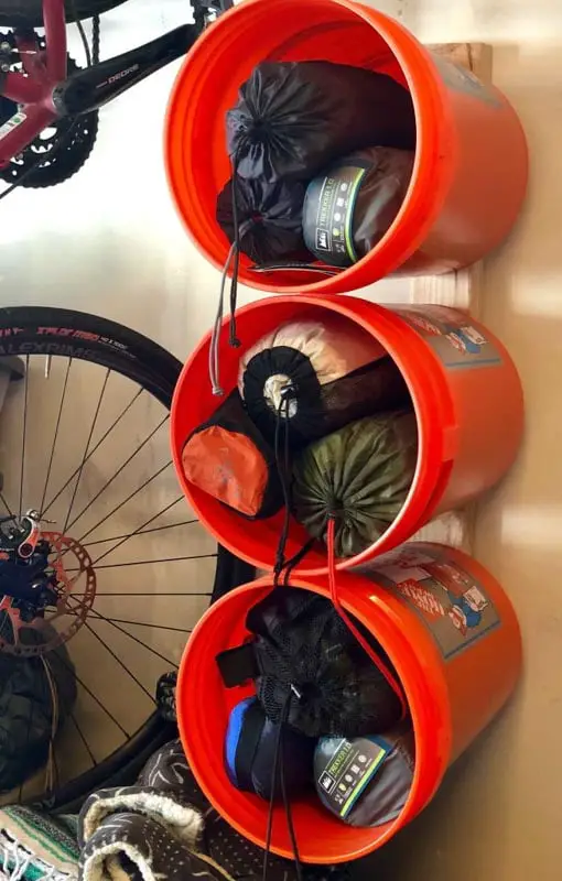 Large, orange paint buckets mounted sideways to a shed wall, holding foldable chairs and sleeping bags.