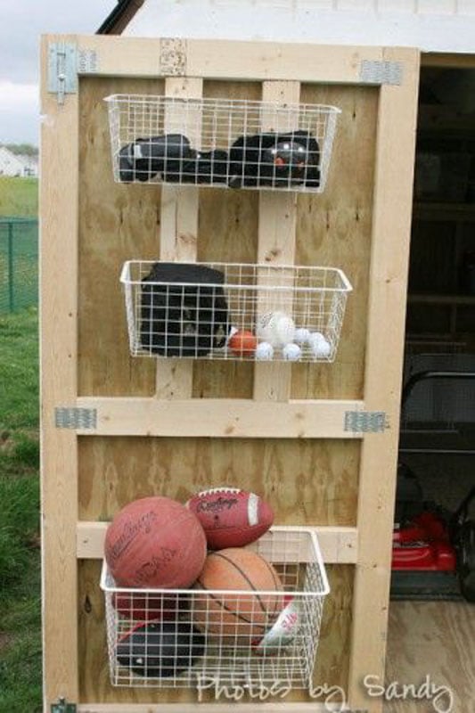 A shed door with white wire baskets mounted inside, filled with sports equipment. 