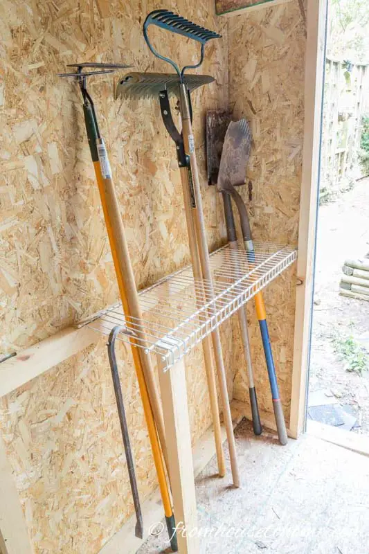 A metal wire shelf affixed to a shed wall for storing long yard tools. 